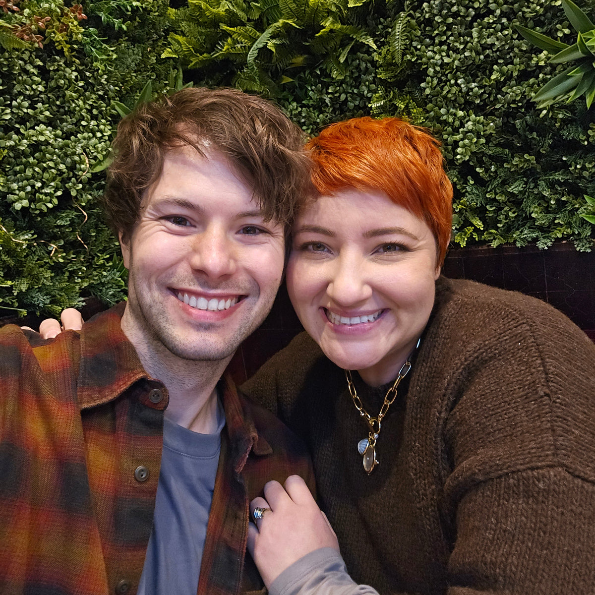 A smiling man and a smiling woman posing together in front of a green foliage wall. The man wears a brown and orange plaid flannel shirt over a gray t-shirt, and the woman has short red hair and wears a brown knit sweater.